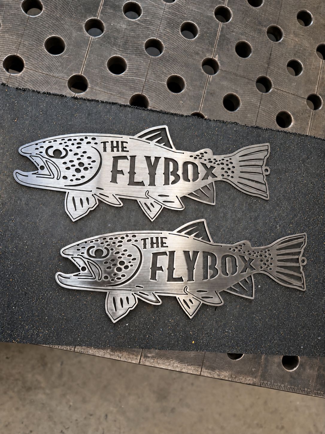 Two custom laser-cut steel fish signs reading The Flybox on the shop table